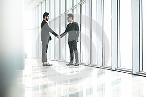 Two indian businessmen shaking hands standing at big window with urban cityscape, confident business partners handshaking in