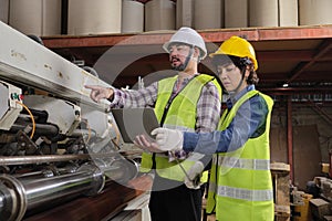 Two industrial workers use a laptop to check a paper manufacturing machine