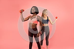 Two international slim girls in sportswear standing one by one, african and europen female`s holding dumbbells in their