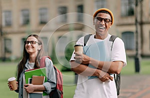 Two international stylish students walk near the campus and smile. Friendly African American guy and Caucasian girl walk