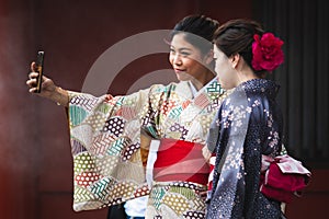 Two Japanese girls taking a selfie with smartphone. Tourists dressed in traditional colorful kimono. Entrance of a shinto temple.