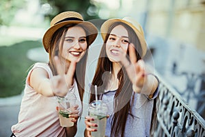 Two joyful attractive girls taking a selfie while walking todether with cocktails on the street and showing peace gesture outdoors