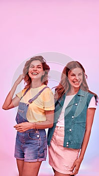 Two joyful attractive young girls, twin sisters posing together isolated over pink background