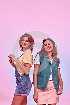 Two joyful attractive young girls, twin sisters smiling, dancing while posing together isolated over pink background