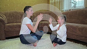 Two joyful Caucasian girls sisters play clapping their hands while sitting on the carpet near the sofa in the living