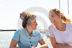 Two joyful girls, friends stand near the shore on the pier and look at each other.