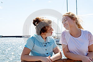 Two joyful girls, friends stand near the shore on the pier and look at each other.