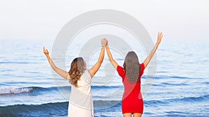 Two joyful girls girlfriends standing on the beach looking at the sea horizon with hands up