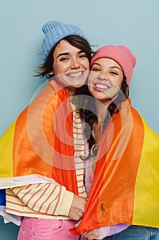 Two joyful girls holding rainbow lgbt flag and hugging isolated over blue background