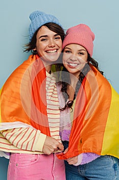 Two joyful girls holding rainbow lgbt flag and hugging isolated over blue background