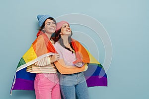 Two joyful girls holding rainbow lgbt flag and hugging  over blue background