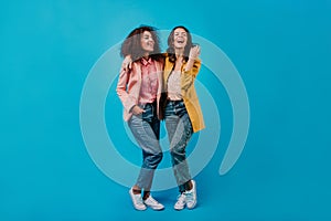 Two joyful girls posing on blue studio background. Full length photoshoot of stylish female models