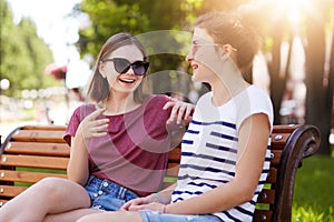 Two joyful girls relax together in local park sitting on new wooden bench, make jokes, gossip, remember old funny moments with