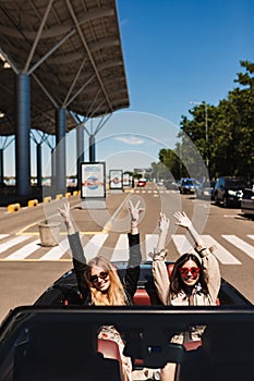 Two joyful girls in sunglasses happily looking in camera while driving cabriolet car on city streets outdoor