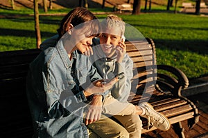 Two joyful girls using smartphone, sitting on the bench in the city park. Lesbian couple holding their phones while