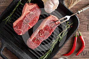 Two juicy, fresh new york steaks in a grill pan with sprigs of rosemary, a head of garlic, pods of red chili pepper, a meat fork