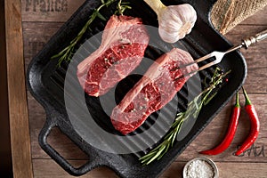 Two juicy, fresh new york steaks in a grill pan with sprigs of rosemary, a head of garlic, pods of red chili pepper, a meat fork