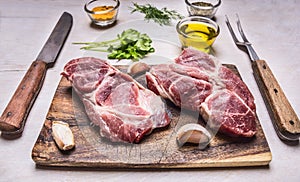 Two juicy fresh raw pork steak on a cutting board with garlic, oil, herbs, a knife and fork on wooden rustic background top view c