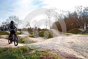 two kids on bicycles ride on a dirt jump track in bright sun under a blue sky.