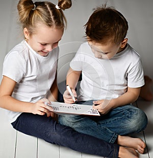 Two kids boy and girl in white t-shirts and blue jeans sit close to each other and play word game with black markers on plate