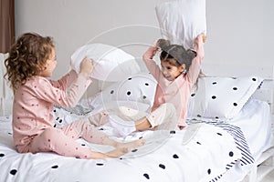 two kids girls in pajamas having pillow fight on bed in modern bright apartment