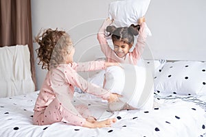 two kids girls in pajamas having pillow fight on bed in modern bright apartment