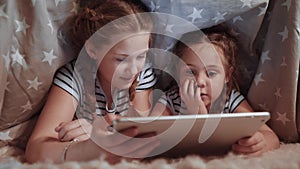 Two kids girls under the blanket with a digital tablet. Kid dream online video games at concept. Daughters kids watching