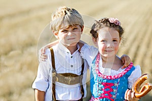 Two kids in traditional Bavarian costumes in wheat field. German children eating bread and pretzel during Oktoberfest