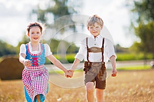 Two kids in traditional Bavarian costumes in wheat field. German children sitting on hay bale during Oktoberfest. Boy