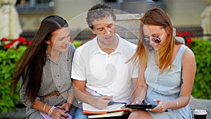 Two Ladies with Charming Smiles and Long Hair Sitting on the Brench near Handsome Boy in White Shirt and Jeans and