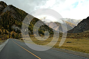 A two-lane asphalt road turns into a wide crevice towards high mountains with peaks in the clouds on a sunny autumn day