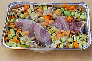 Two large pieces of back of pork, chopped mushrooms and vegetables in a foil container