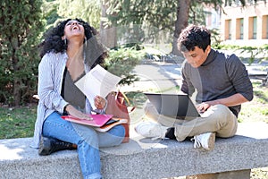 Two latin students laughing studying together sitting on a bench outdoors