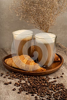 Two lattes with foam tops beside a golden croissant on a wooden tray, surrounded by coffee beans photo