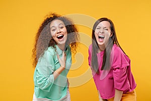Two laughing excited women friends european and african american girls in pink green clothes posing isolated on yellow