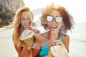 Two laughing friends drinking from coconuts on a sandy beach