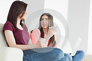 Two laughing girls sitting on a sofa enjoying a beverage and holding a laptop