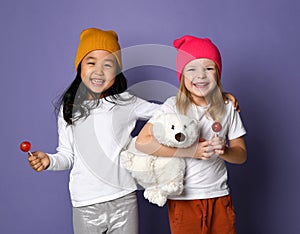 Two laughing kid girls in white t-shirt and colorful hats hold polar bear toy and lollipop candy on purple