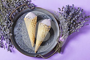 Two lavender ice creams in waffle cones on a tin tray and bouquets of dry lavender on a lilac background.