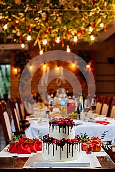 Two level white wedding cake, decorated with fresh red fruits and berries, drenched in chocolate. Bright banquet table decoration