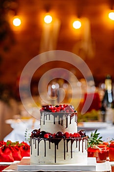 Two level white wedding cake, decorated with fresh red fruits and berries, drenched in chocolate. Bright banquet table decoration