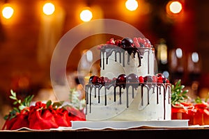 Two level white wedding cake, decorated with fresh red fruits and berries, drenched in chocolate. Bright banquet table decoration