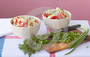 Two light bowls with a salad of fresh vegetables are on the table covered with a tablecloth, next to a cutting board, a knife,