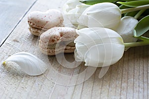Two light tasty creamy macarons are lying on the wooden table with white tulips. Two macarons in form of the heart as 8