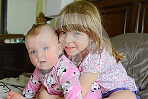 Two little adorable Caucasian sisters sit together.The little sister hugs her baby-sister. Girls are smiling and happy