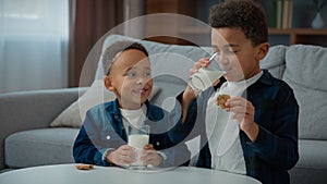 Two little African American boys kids ethnic adopted hungry children drink milk eat cookies at home living room siblings