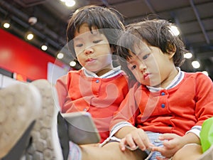 Two little Asian baby girl, sisters, sitting and watching a smartphone together, while waiting for her mother to do some errands photo
