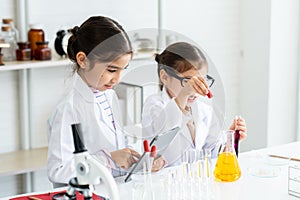 Two little Asian girls in white lab coat help each other for experiment. Little glasses girl using pipette drop solution in yellow