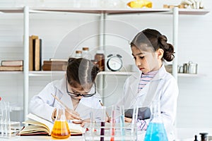 Two little Asian girls in white lab coat help each other for experiment. Little glasses girl writing her new idea by pencil for