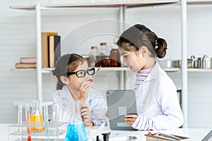 Two little Asian girls in white lab coat help each other for experiment. Little glasses girl showing violet solution inside